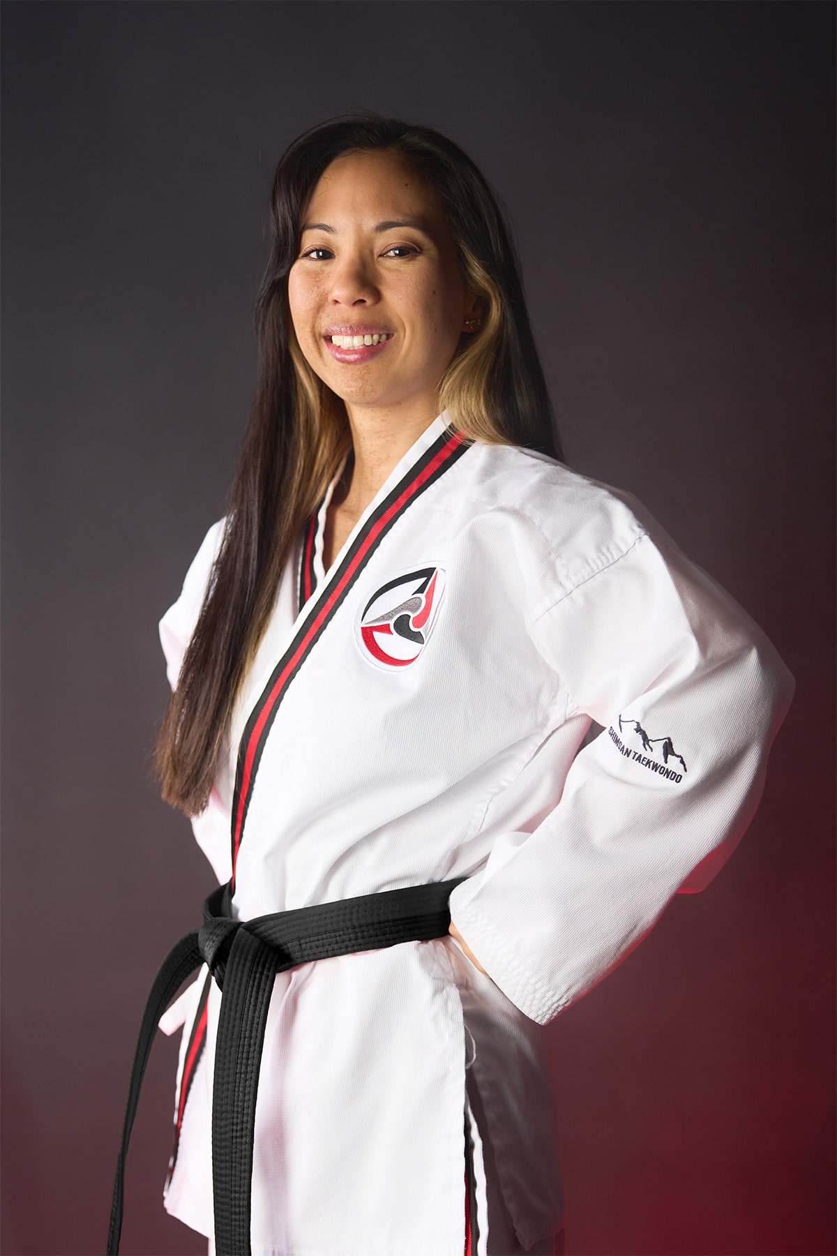 Ms. Janel Cancino wears a white taekwondo uniform and a black belt, with her hands on her hips.